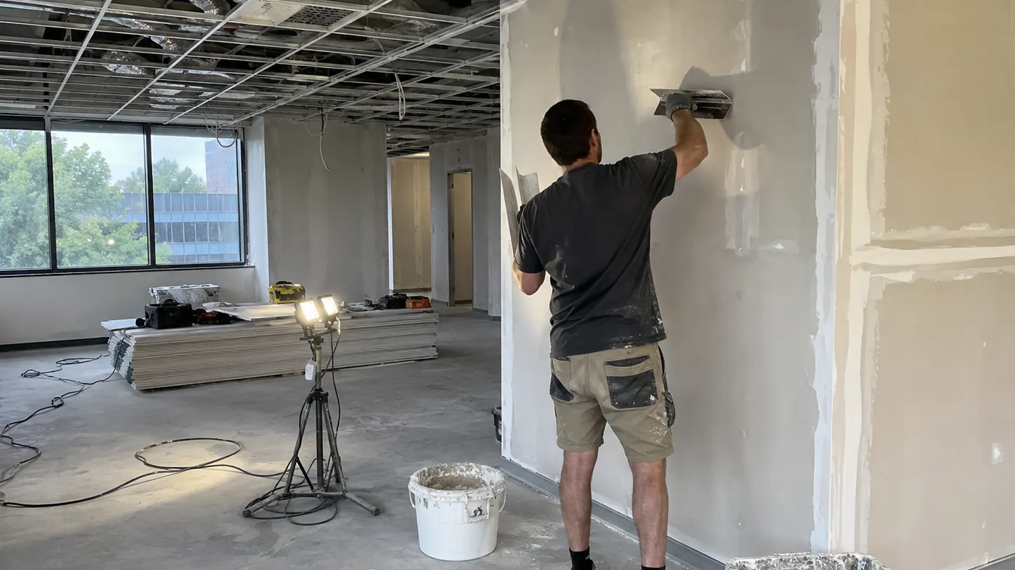 Commercial plasterer finishing an office wall during a commercial fit-out with exposed ceiling grid and work equipment.